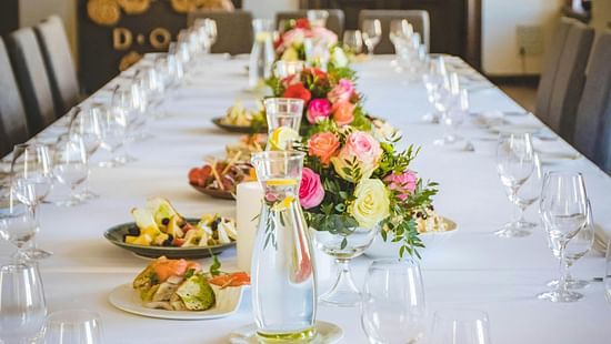 A long dining table decorated with flowers in the middle along with shiny glassware and cutlery set on the table.