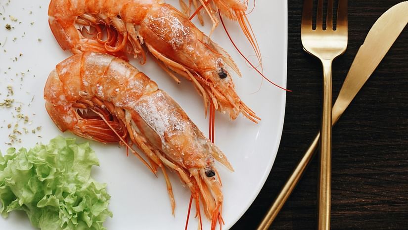 A white plate of cooked shrimp, garnished with parsley, sits on a dark wooden table next to a fork and knife.