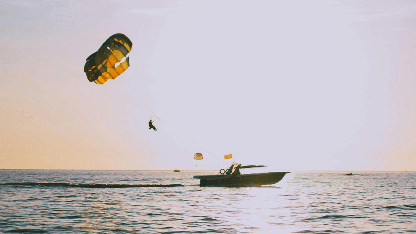 A person parasailing with a colorful parachute over calm sea waters during golden hour with a motorboat visible on the water surface and a soft pastel sky in the background.