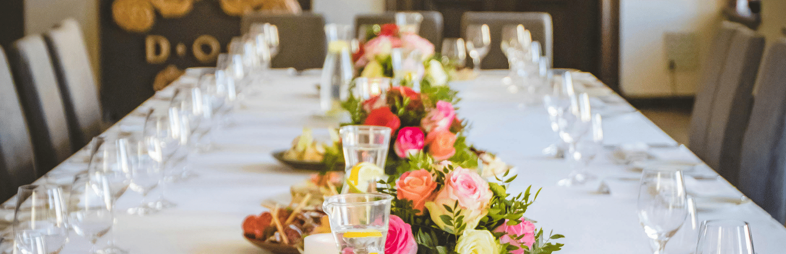 A long dining table decorated with flowers in the middle along with shiny glassware and cutlery set on the table.