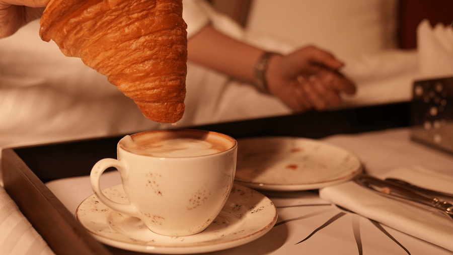 A person in a white robe holds a large croissant above a cup of frothy coffee on a breakfast tray, creating a cosy, relaxed morning ambiance.