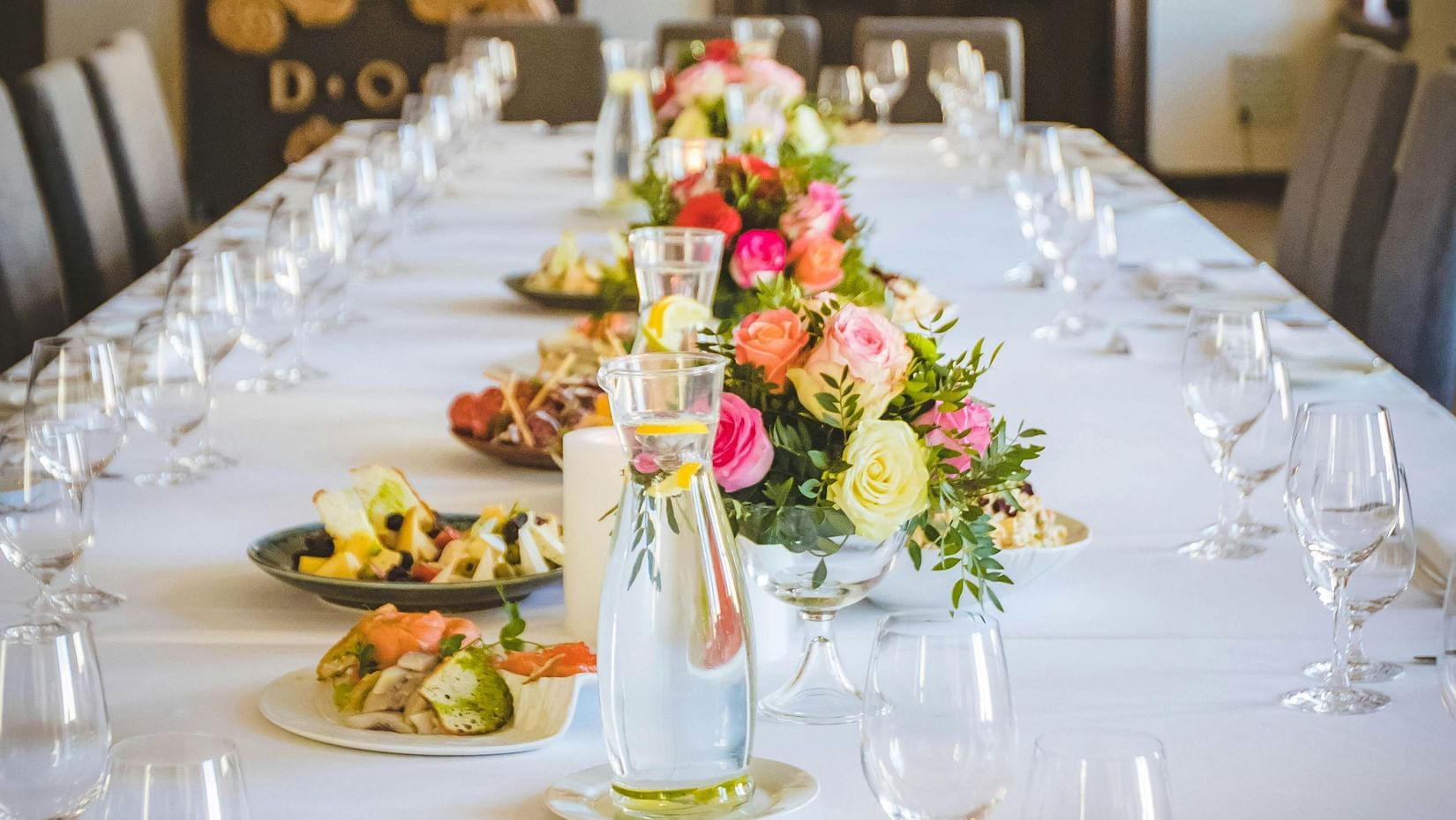 A long dining table decorated with flowers in the middle along with shiny glassware and cutlery set on the table.