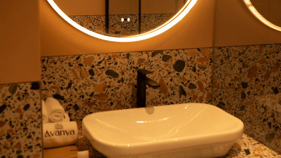 A white sink on a terrazzo counter with illuminated round mirror and a reflection of another at Avanya Resorts N Retreat.