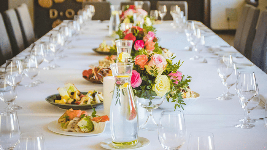A long dining table decorated with flowers in the middle along with shiny glassware and cutlery set on the table.