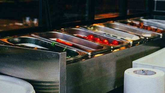 A close up of a buffet station with trays of food, stacked plates, and paper towels