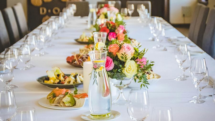 A long dining table decorated with flowers in the middle along with shiny glassware and cutlery set on the table.