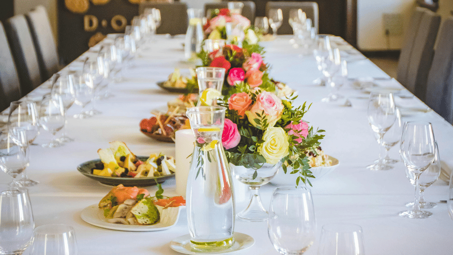 A long dining table decorated with flowers in the middle along with shiny glassware and cutlery set on the table.