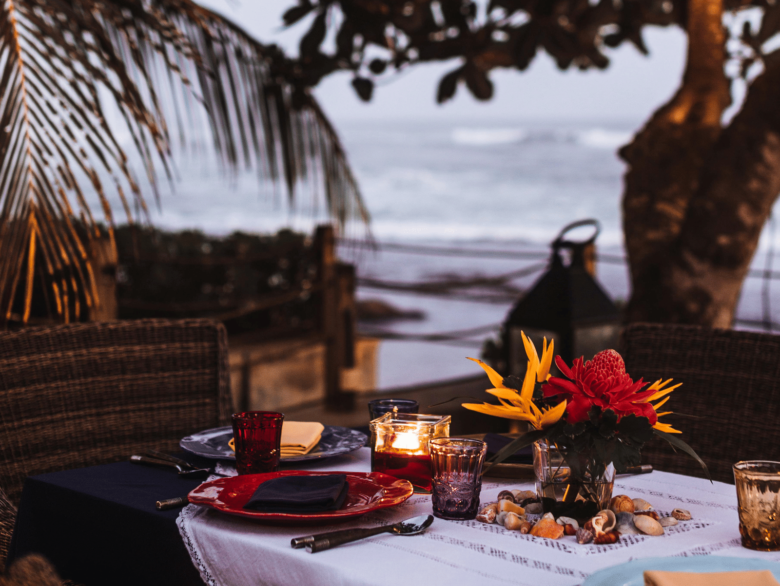 A cosy beachside dining setup under palm trees with a table laid out for two, offering a peaceful tropical ambience with ocean views.