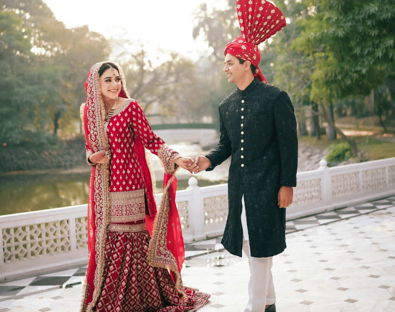 The bride and groom, dressed in rich traditional outfits, walk hand-in-hand along a paved path next to a body of water in soft daylight.