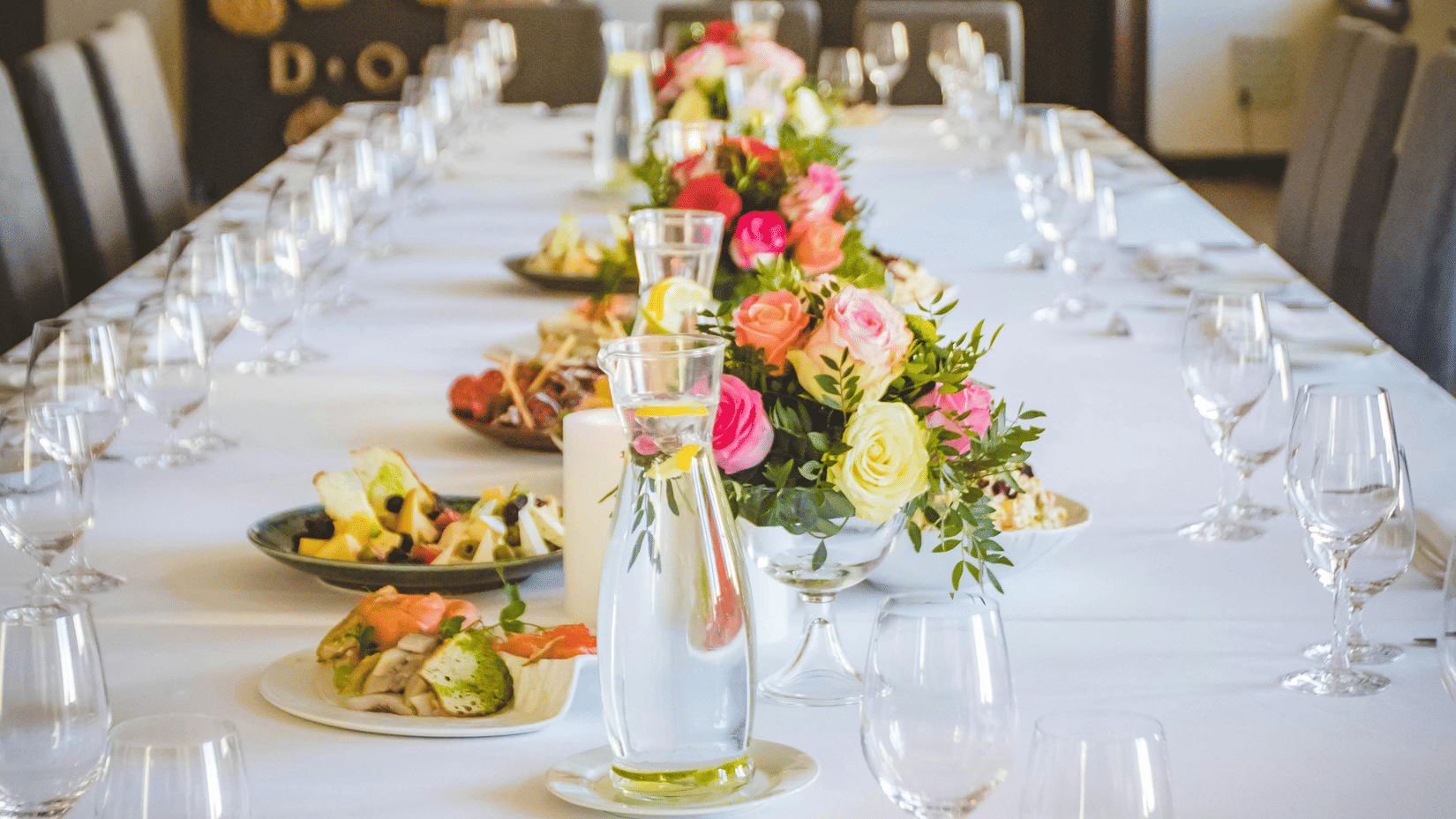 A long dining table decorated with flowers in the middle along with shiny glassware and cutlery set on the table.