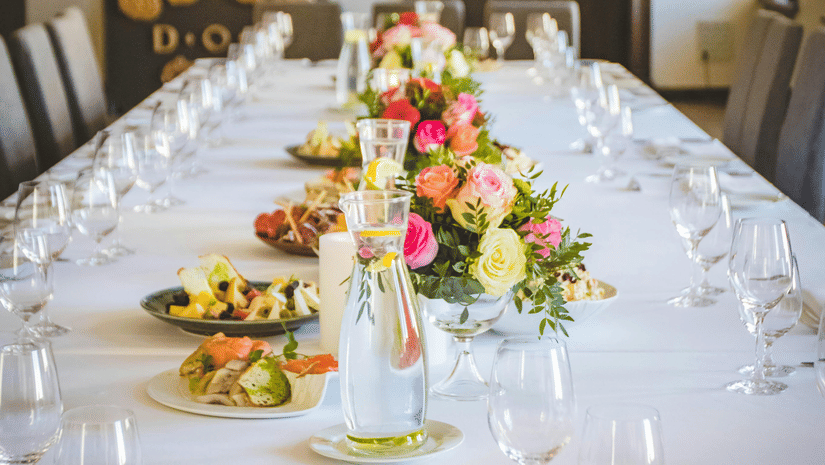 A long dining table decorated with flowers in the middle along with shiny glassware and cutlery set on the table.