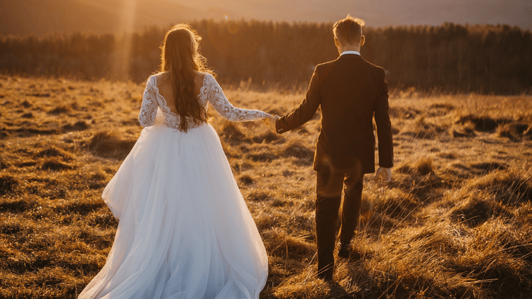 A newlywed couple walks hand-in-hand across a sunlit field with mountains in the distance