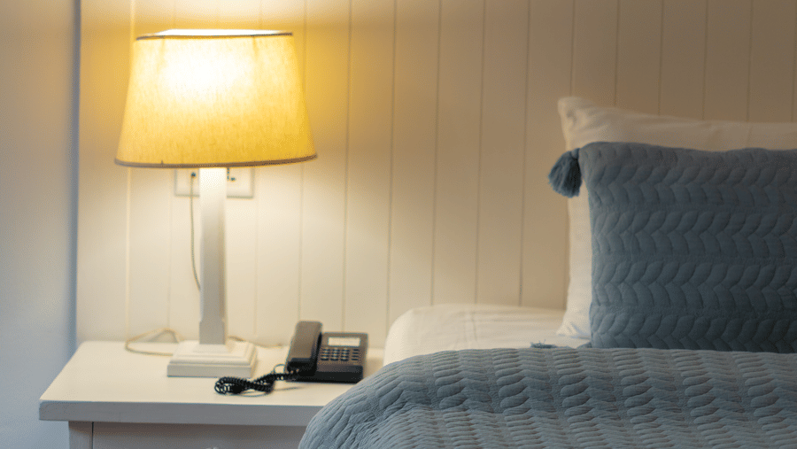 A bedside setup with a warm white lamp on a white nightstand next to a bed with light blue linens - Johnson Lodge & Spa, Manali