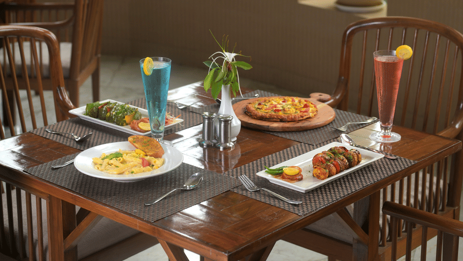 Restaurant table arranged with multiple plated dishes, cutlery and glassware beside wooden dining chairs and large windows.