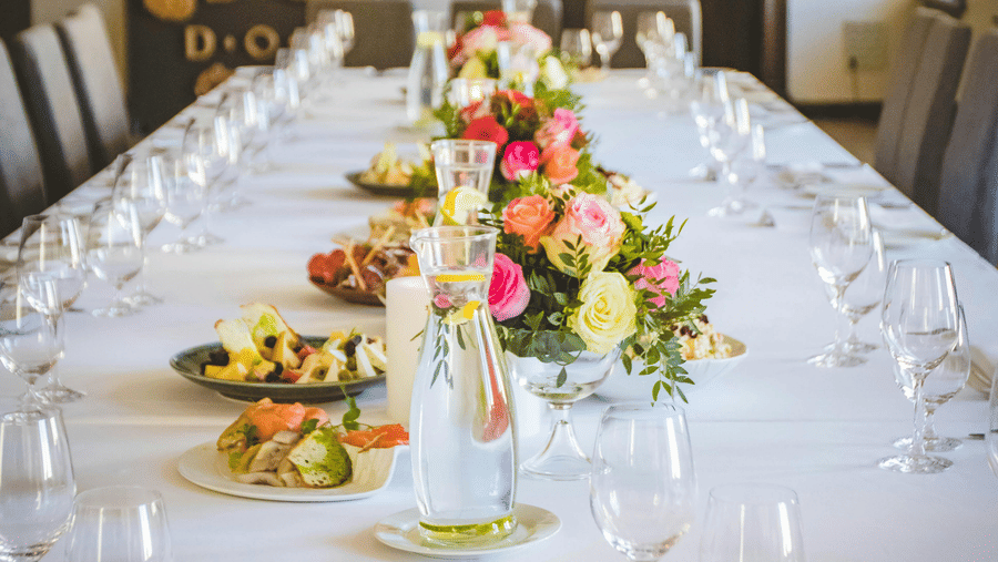 A long dining table decorated with flowers in the middle along with shiny glassware and cutlery set on the table.