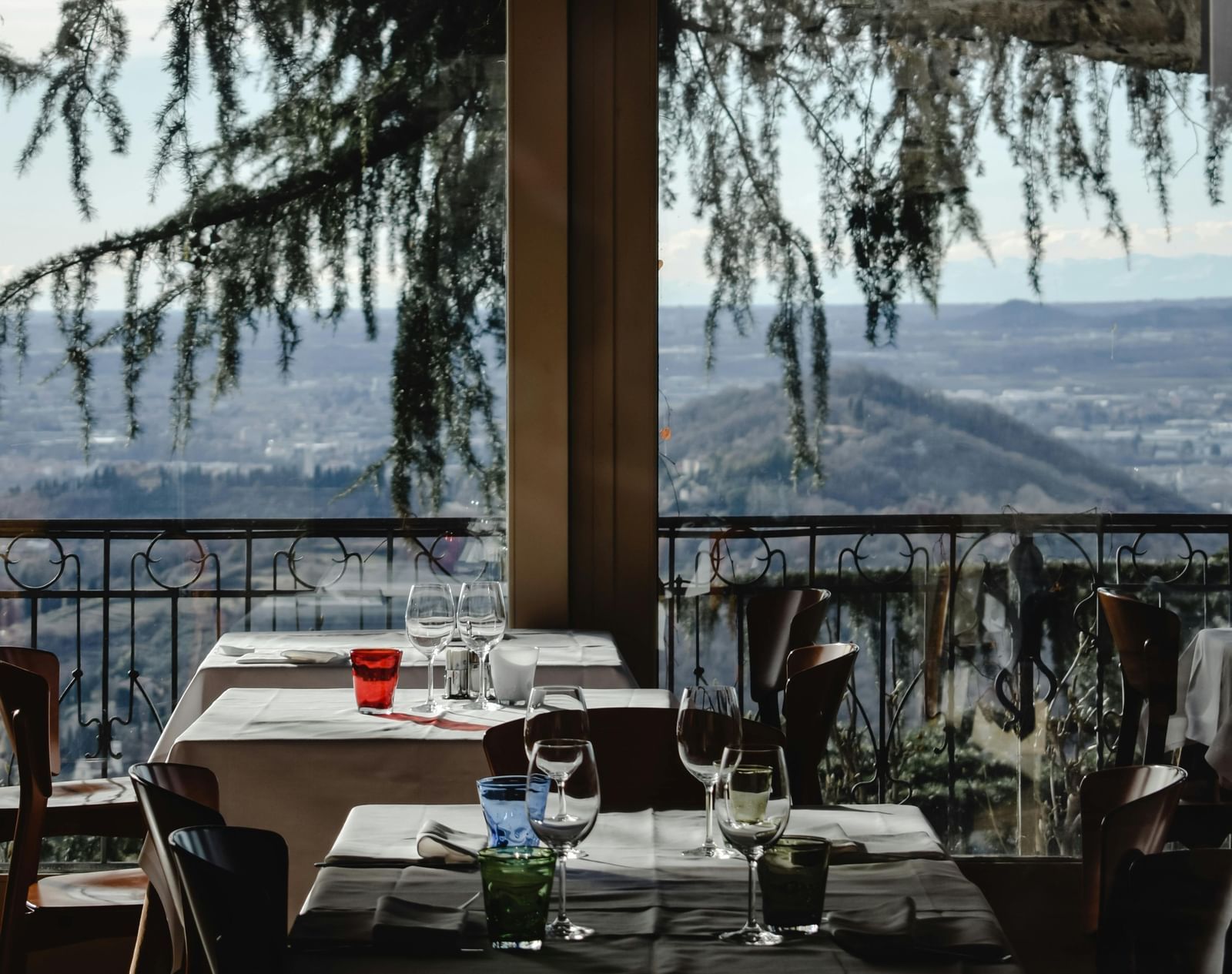 Indoor restaurant dining area with set tables, glassware, and a scenic view of mountains and trees visible through large windows in the background.