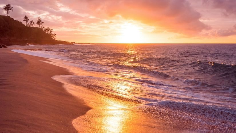 The shore of a beach during sunset, with waves reflecting the warm colours of the sky.
