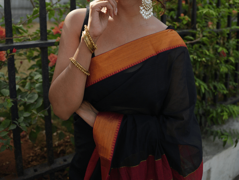 A woman posing for a picture while wearing a sleeveless blouse and one of the best high-quality Chettinad cotton sarees, standing in front of a building with trees in the background.