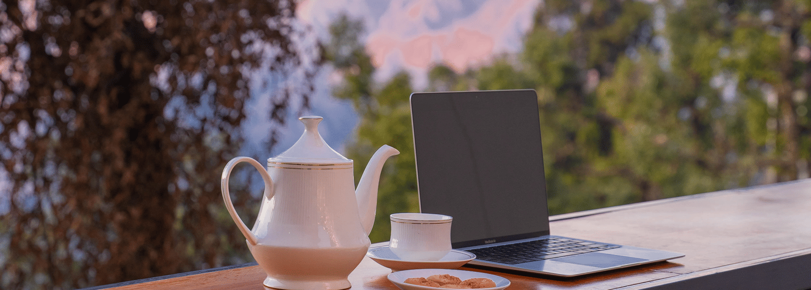 Close-up shot of a laptop along with a tea pot and a cup at Perfectstayz Premium Westend Mall Road, Mussoorie, with a beautiful backdrop of the mountains.