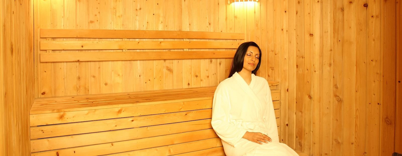 A woman in a bathrobe sitting on a wooden bench, enjoying a steam bath, surrounded by a misty atmosphere that enhances relaxation