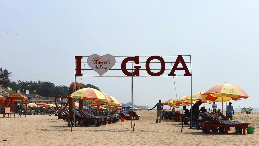 'I Love Goa' sign stands on a sandy beach under a clear sky, with colourful sun umbrellas and loungers behind.
