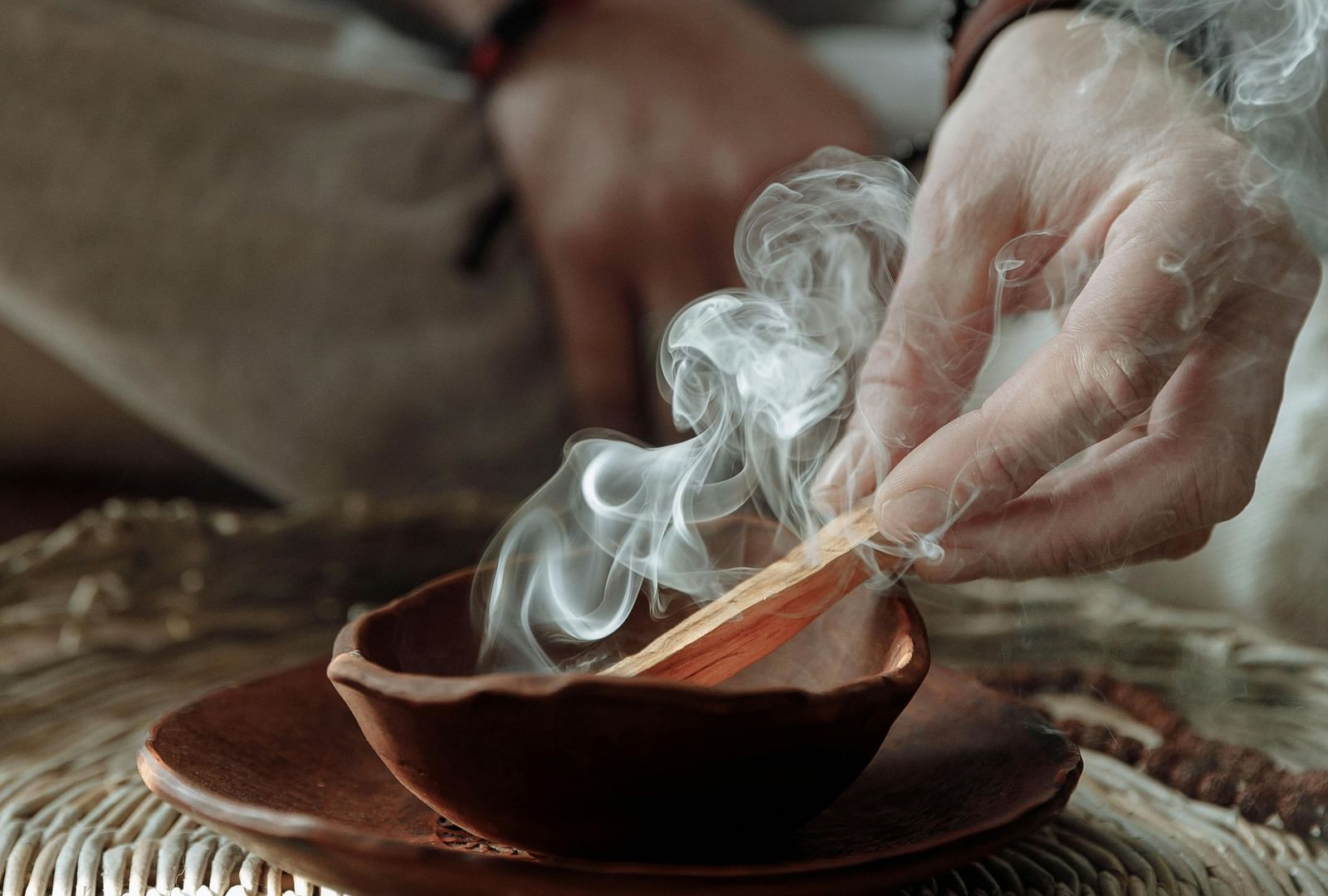 A person sitting on a huge mat burning incense sticks into a bowl.
