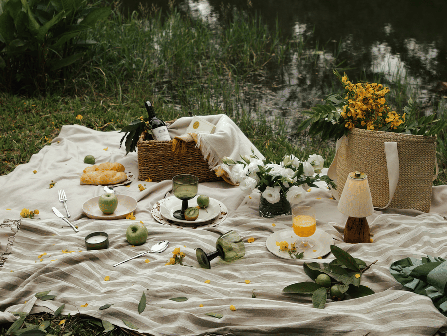 A luxurious lakeside breakfast picnic at The Serai Chikmagalur, set on a striped blanket with fresh pastries, fruits, juice, and elegant floral touches amid lush greenery.