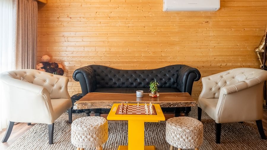 A warmly lit lounge area with wooden walls and ceiling features a black sofa, two cream armchairs, a bright yellow coffee table, two small stools, and a ceiling fan at Themis Farm House.