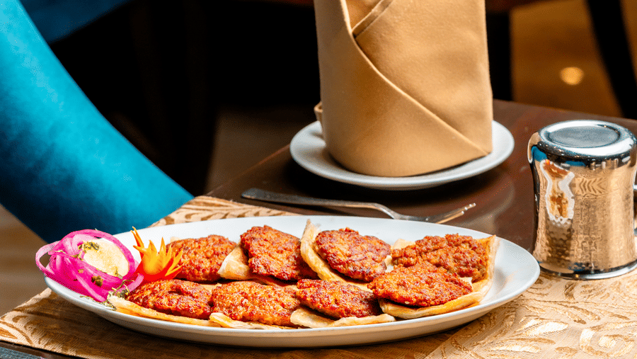 A platter of reshmi kebabs served at a table in Za-aiqa, Noor-Us-Sabah Palace, Bhopal, garnished with sliced onions and lime, with a folded tan napkin and teal-upholstered chair visible.