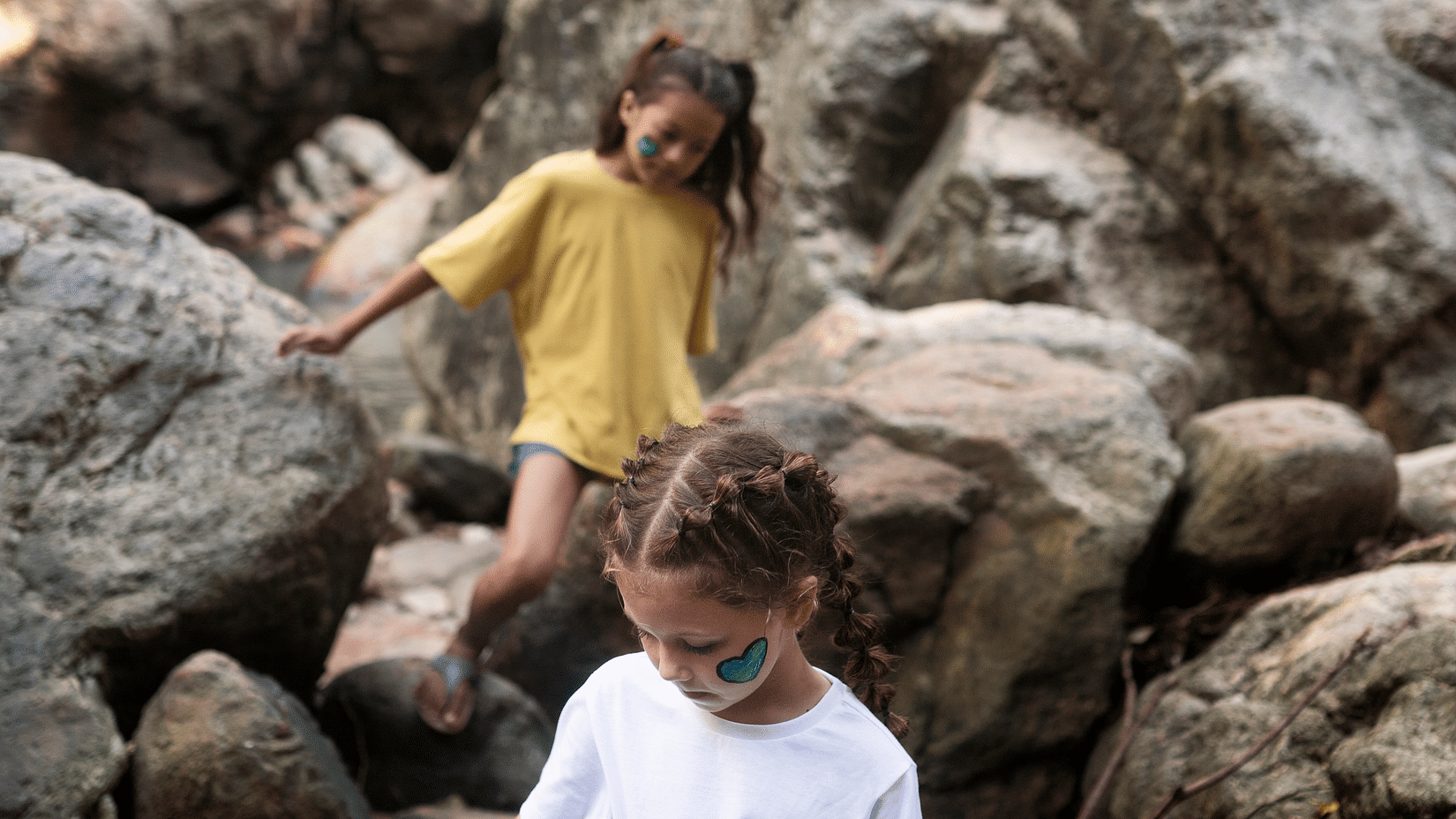 Three children explore a rocky forest area, climbing and balancing on stones with painted faces.