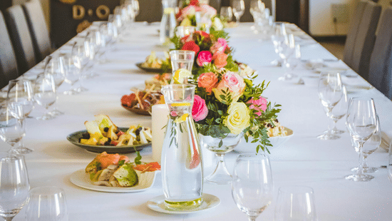 A long dining table decorated with flowers in the middle along with shiny glassware and cutlery set on the table.