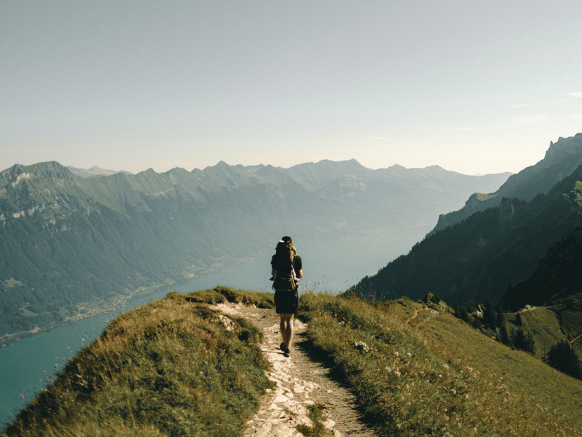  A solitary hiker with a backpack walks away from the camera on a grassy, dirt path along a ridge overlooking a vast valley and mountain range under a pale sky.