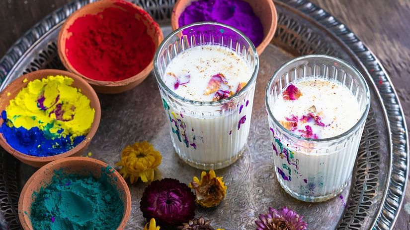 Two glasses of Thandai, a traditional Indian drink, garnished with flower petals, sit on a tray with colorful Holi powders.