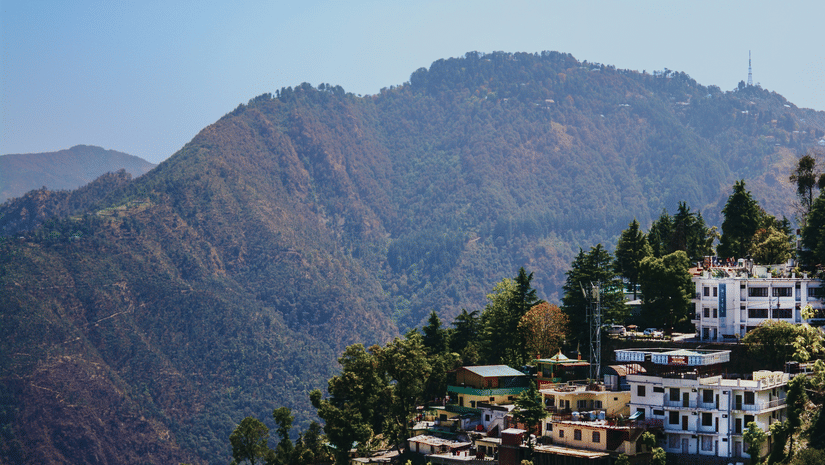 Mountains of Mussoorie featuring houses on them under a clear sky on a sunny day with surrouding lush trees