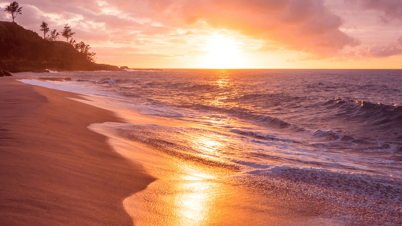The shore of a beach during sunset, with waves reflecting the warm colours of the sky.
