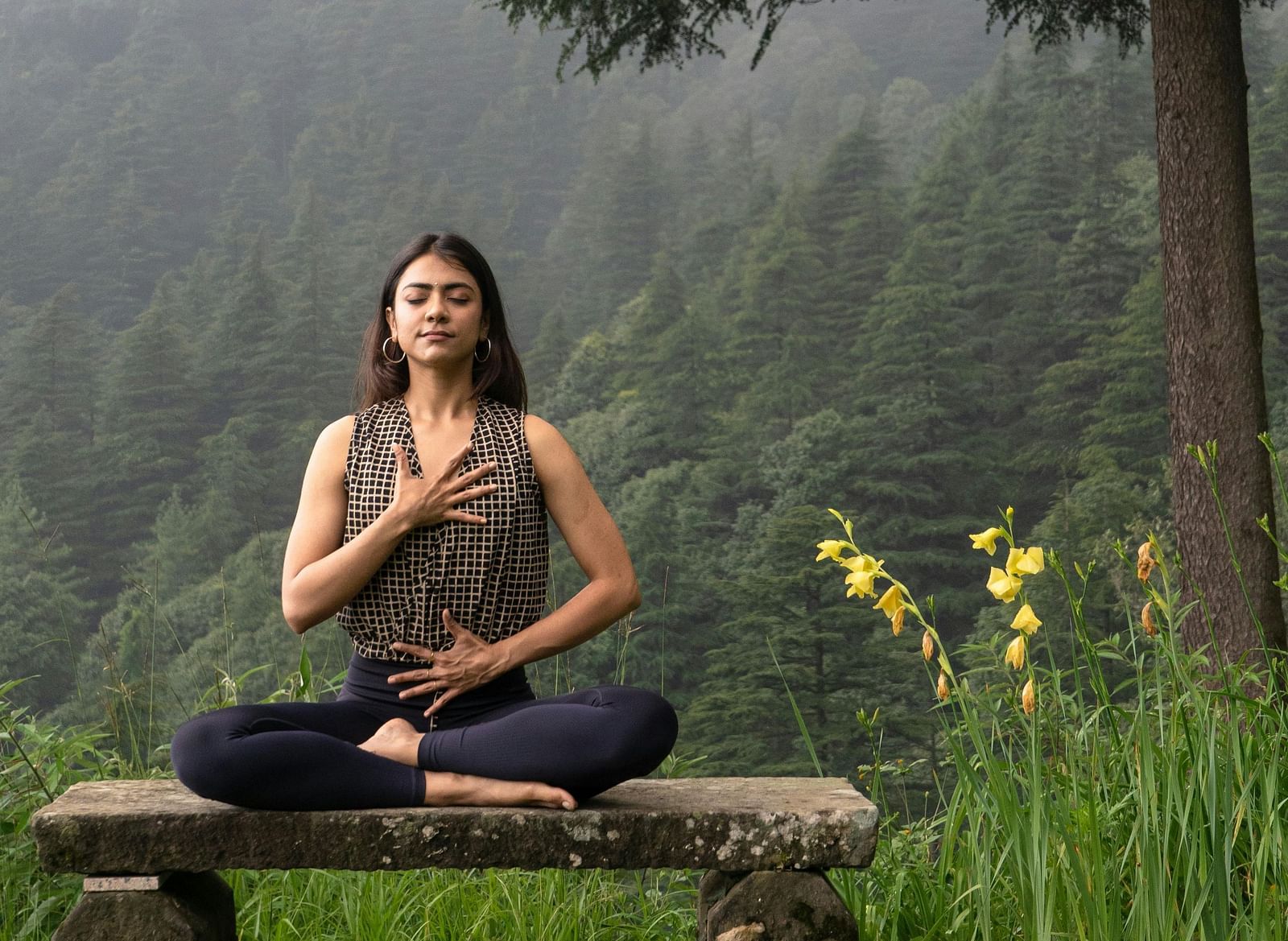 A woman sitting on a low height bench meditating with a misty valley behind her.