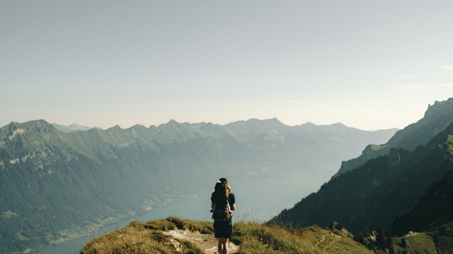  A solitary hiker with a backpack walks away from the camera on a grassy, dirt path along a ridge overlooking a vast valley and mountain range under a pale sky.