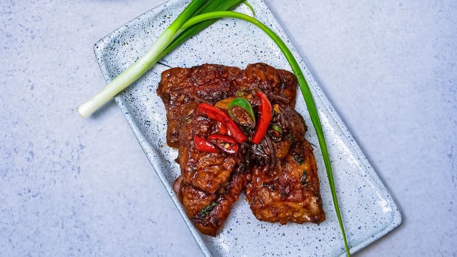 A rectangular glass plate with a grilled dish garnished with green onions, served beside a bowl of vegetable soup on a light blue textured background at The Hamuse Hotel, Kodaikanal.