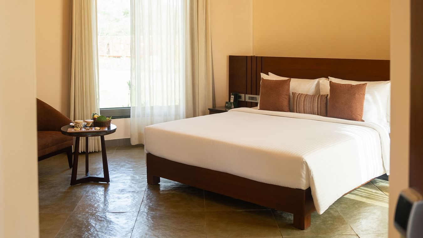 A cosy bedroom with a double bed, arched ceiling, ceiling fan, seating space and natural light through a large window at Ananta Spa and Resort, Ajabgarh.