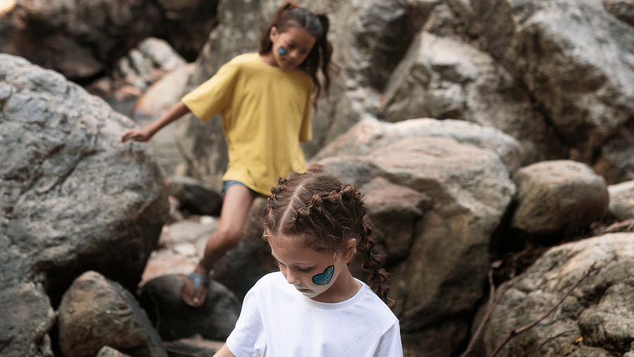 Three children explore a rocky forest area, climbing and balancing on stones with painted faces.