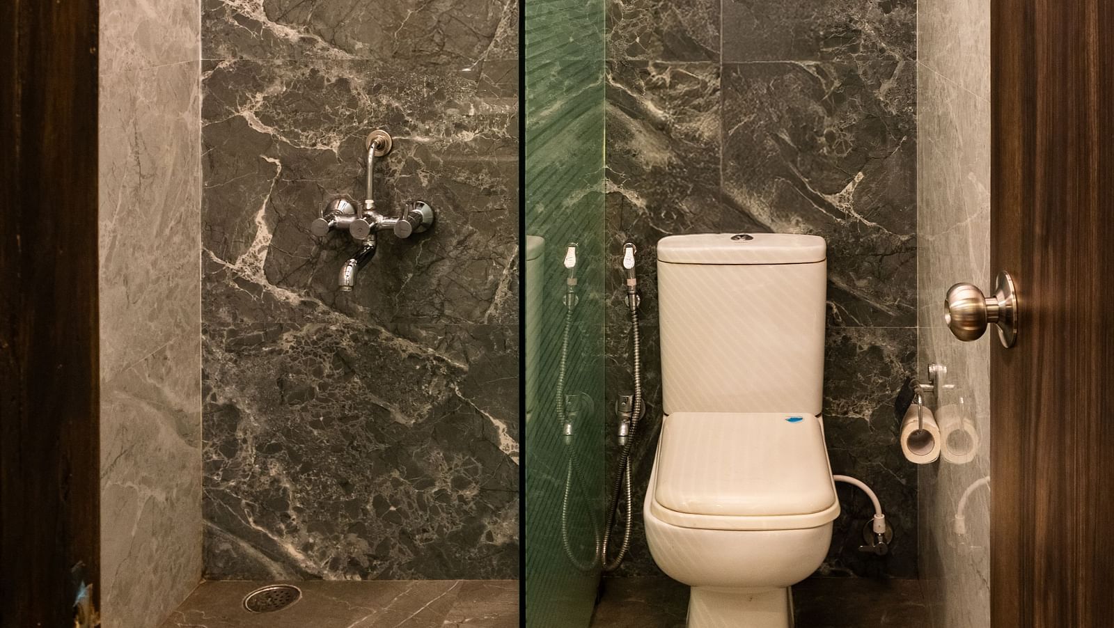 Compact and stylish bathroom with marble-textured walls and flooring, a glass partition for the shower area, and a Western toilet under a designer ceiling - The Grand Legacy Resort & Spa