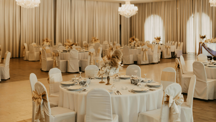 A banquet hall with round tables, chairs covered in fabric, ceiling lights, and a stage area at the far end.