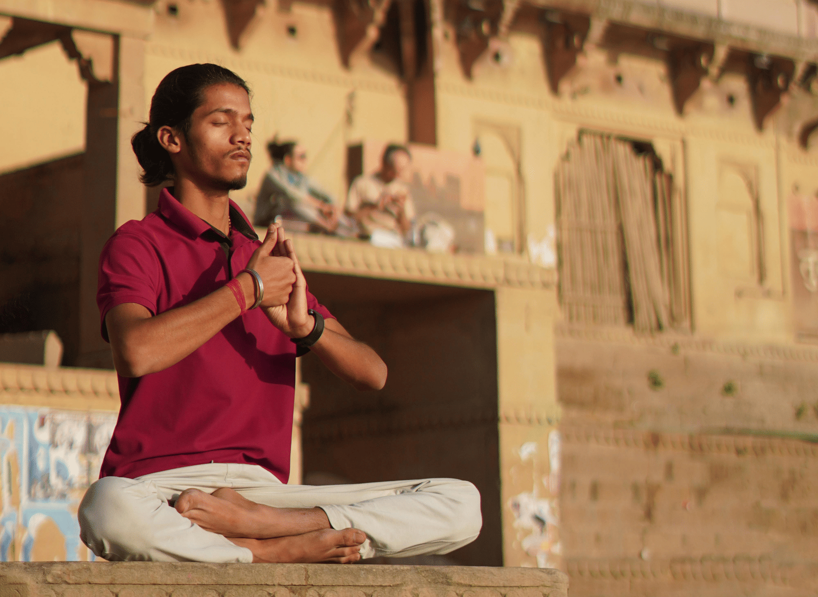 A man in yoga posture, meditating on a stone bench-like structure, with traditional building with Indian architecture in the background.