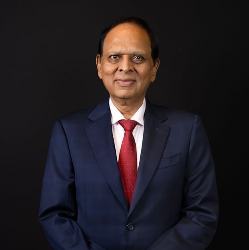 Professional portrait of a man in a navy suit with a red tie, standing with hands clasped against a black background.