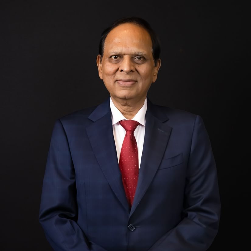 Professional portrait of a man in a navy suit with a red tie, standing with hands clasped against a black background.