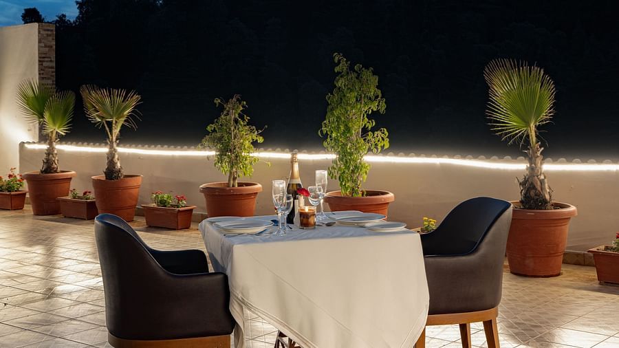An outdoor dining setup at Clarks Inn, Near Kainchi Dham at dusk features a set table for two, surrounded by potted plants and soft string lighting.