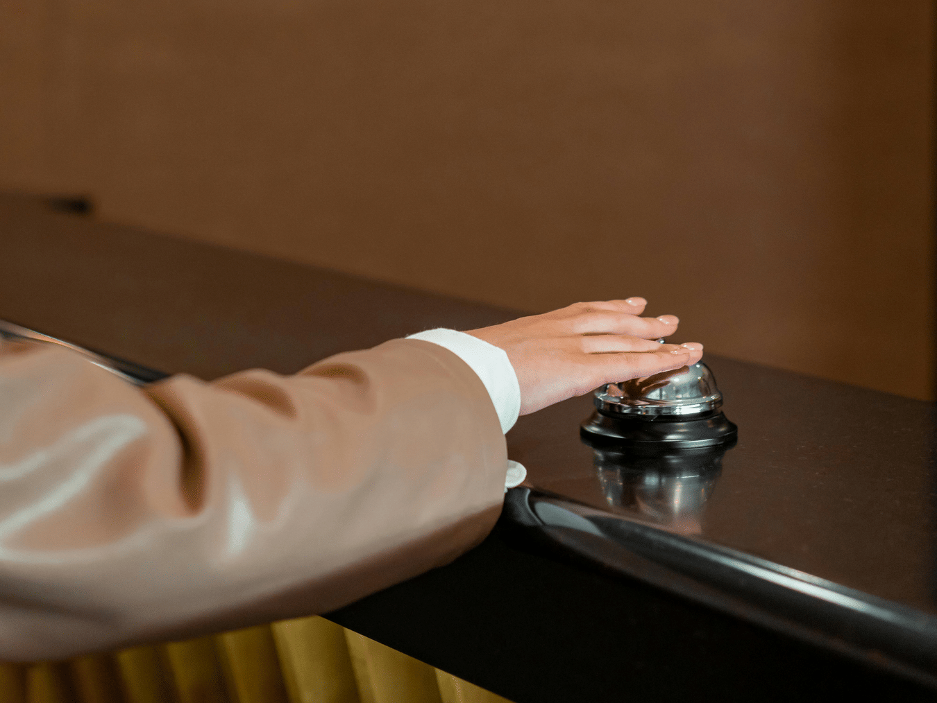 Person pressing a silver service bell on a wooden front desk counter.