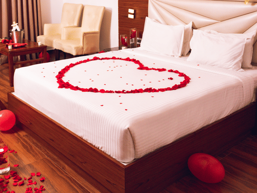 A hotel room decorated for a romantic occasion, with a heart-shaped rose petal arrangement on the bed and love balloons.