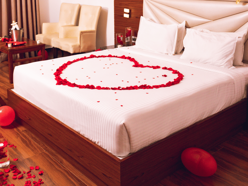 A hotel room decorated for a romantic occasion, with a heart-shaped rose petal arrangement on the bed and love balloons.