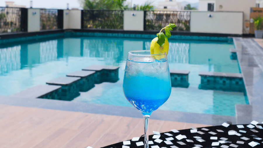 A single cocktail glass on a patterned ledge overlooking an outdoor pool and distant buildings | Pride Premier Alwar
